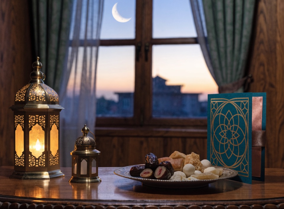 An Eid greeting card beside glowing lanterns, dates and a crescent moon in the window